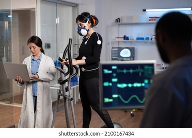 Athlete Sportwoman With Electrodes Attached To Her Body Running On Cross Trainer Doing Cardio Exercise. Specialist Researcher Holding Laptop Analyzing Endurance Controling EGK Data In Modern Lab