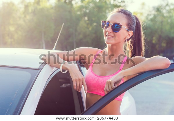 Athlete\
sporty fit young woman in sports bra wearing sunglasses standing\
leaning on car with door open looking at\
camera
