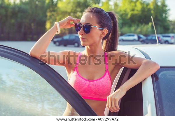 Athlete\
sporty fit young woman in sports bra wearing sunglasses standing\
leaning on car with door open looking at\
camera.