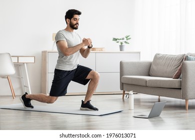Athlete trains at home, new normal and online lesson during covid-19. Cheerful arabian bearded young guy doing lunges, for legs in living room interior, looks at laptop on floor, near bottle of water