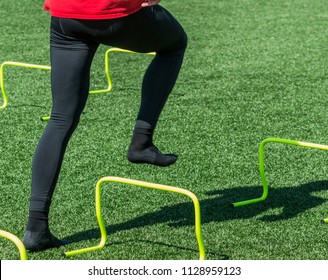 An Athlete Wearing A Red Shirt Abd Black Spandex Is Stepping Over Yellow Mini Banana Hurdles Wearing Socks On A Green Turf Field.