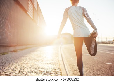 Athlete woman preparing for running on the city street. Legs warming and stretching. Sport tight clothes. Bright sun, blurry background. Horizontal