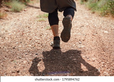 Athletes Runner Running Down A Mountain Trail. A Man Runs Through The Mountains Between The Stones. View From Behind. Man In Shorts And Black Spandex Leggings Training Outdoors.