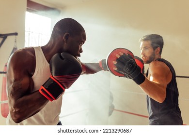 Athletic African Man Hitting The Focus Mitts Held By His Personal Trainer In A Gym. Young Boxer Having A Training Session With His Coach.