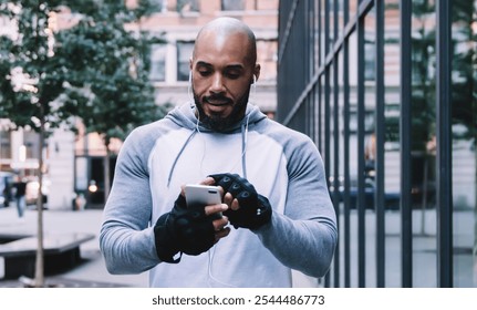 Athletic bald bearded African American sportsman listening to music in earphones and browsing smartphone while standing on pavement near modern office building - Powered by Shutterstock