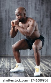 Athletic bare-chested African American doing squats while holding hands near chest in gym.