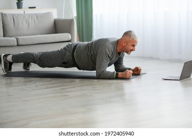 Athletic Bearded Older Man Doing Exercises At Home, Using Laptop, Watching Sport Videos Online And Planking, Having Fitness Class Online, Copy Space. Healthy Lifestyle, Sport On Self-isolation