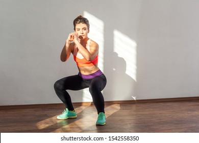 Athletic Beautiful Woman With Bun Hairstyle And In Tight Sportswear Practicing, Squatting Doing Sit-up At Home Or Fitness Gym, Concentrated Look. Indoor Studio Shot Illuminated By Sunlight From Window