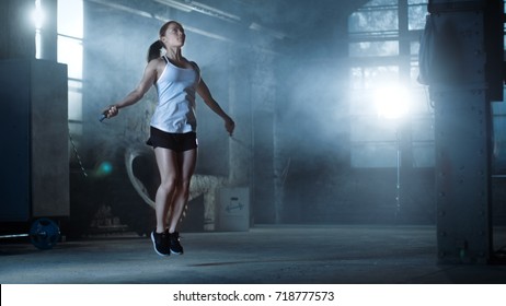 Athletic Beautiful Woman Exercises with Jump / Skipping Rope in a Gym. She's Covered in Sweat from Her Intense Fitness Training. Dark atmosphere.