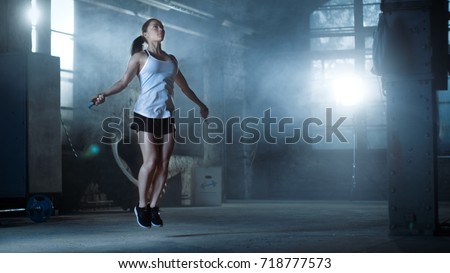Athletic Beautiful Woman Exercises with Jump / Skipping Rope in a Gym. She's Covered in Sweat from Her Intense Fitness Training. Dark atmosphere.
