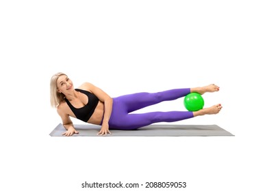 Athletic beautiful woman lying on side on a mat with a small pilates ball between her feet, legs up, isolated on white.