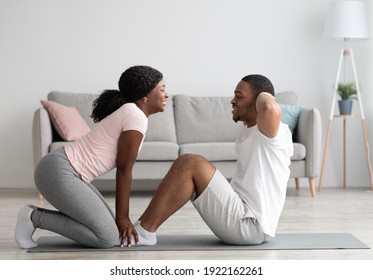 Athletic black couple exercising together at home, working on abs. Cheerful african american woman in sportswear holding her man legs while he is doing exercises for belly, having sport together