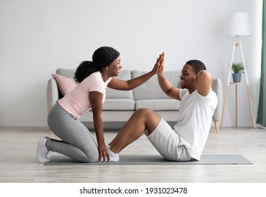 Athletic black couple exercising together at home, working on abs. Cheerful african american woman in sportswear holding her man legs while he is doing exercises for belly, cheering with high five - Powered by Shutterstock