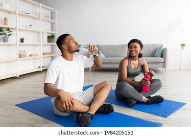 Athletic black couple in sportswear sitting on yoga mats, drinking water from bottles after domestic training, indoors. Cool guy and his girlfriend taking break after workout, staying hydrated