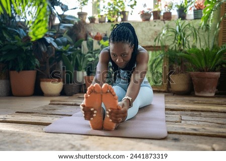 Athletic black woman in sportswear doing limbering-up fitness stretching exercises at home in urban jungle space. Focused african american female warm up muscles before workout sitting on sports mat.
