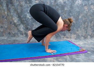 An athletic brown haired woman is balancing in  yoga exercise posture Bakasana or crane crow pose on yoga mat in studio with mottled background.