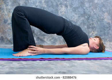 an athletic brown haired woman is doing yoga exercise  Setu Bandhasanasana or advanced bridge pose on in studio against mottled background.
