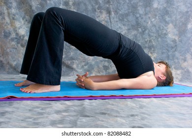 an athletic brown haired woman is doing yoga exercise  Setu Bandhasanasana or bridge pose on in studio against mottled background.