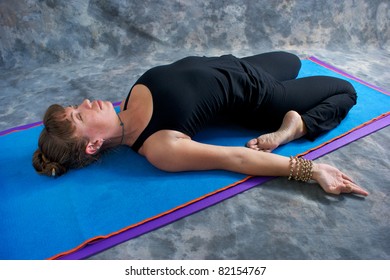 An athletic brown haired woman is doing yoga exercise Reclined hero posture on yoga mat in studio with mottled background.