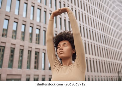 Athletic calm woman with curly hair raises arms over head does stretching exercises wears beige tracksuit breathes deeply poses against modern building leads healthy lifestyle. Wellness concept - Powered by Shutterstock