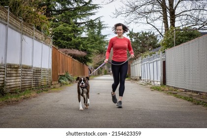 Athletic Caucasian Adult Woman Running Outside With A Boxer Dog. Suburban Neighborhood In A Modern City Of Fraser Heights, Surrey, Vancouver, British Columbia, Canada.