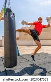 Athletic Caucasian Man Exercising With Black Punching Bag, Having Workout At Street Gym Yard. Sports And Health Concept. Outdoor Training Concept. Selective Focus. Vertical Shot