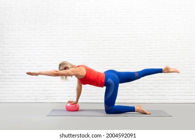 Athletic caucasian woman practice pilates with props in fitness studio indoor, bird dog drill with one leg and arm up and a small fit ball under her hand, selective focus.