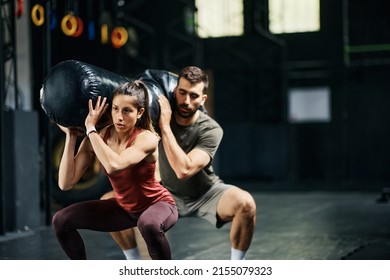 Athletic Couple Doing Squats While Holding Sandbag On Shoulders During Gym Workout. Focus Is On Woman. 