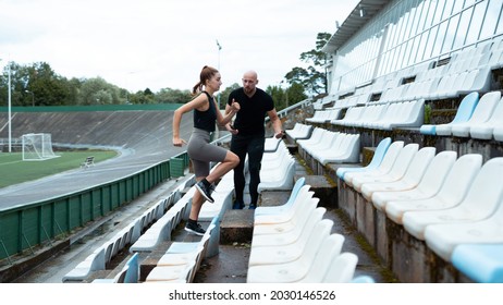 Athletic Couple Working Out Together Outside On A Green Stadium Field. Wearing Dark Workout Clothes, Bright Blue And Black Sneakers. Man Motivating Woman To Run Faster.