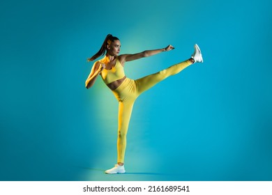 Athletic Female Doing Karate Side Kick Clenching Fists And Raising Leg Standing Training Over Blue Studio Background. Marital Arts And Sport. Self Defense For Women Concept. Full Length