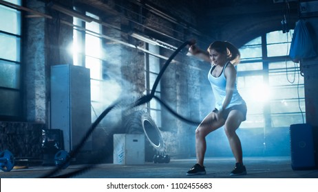 Athletic Female in a Gym Exercises with Battle Ropes During Her Cross Fitness Workout/ High-Intensity Interval Training. She's Muscular and Sweaty, Gym is in Deserted Factory. Cold Ambient. - Shutterstock ID 1102455683