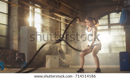 Athletic Female in a Gym Exercises with Battle Ropes During Her Fitness Workout/ High-Intensity Interval Training. She's Muscular and Sweaty, Gym is in Deserted Factory.