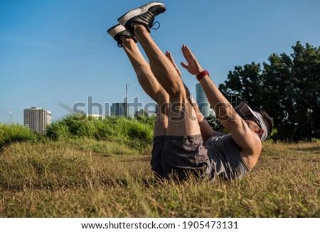 An athletic and fit asian guy does V-ups on the grass at an open field near the city. Abdominal workout or calisthenics outdoors..