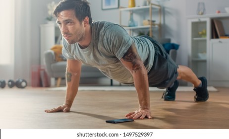 Athletic Fit Man in T-shirt and Shorts is Doing Push Up Exercises While Using a Stopwatch on His Phone. He is Training at Home in His Living Room with Minimalistic Interior. - Powered by Shutterstock