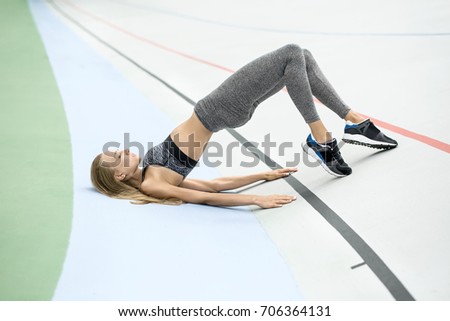 Athletic girl with closed eyes in gray sportswear does stretching exercise on the cycle track outdoors. She holds her feet on the toes and raises her pelvis while relies on the hands and shoulders.