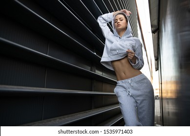 Athletic girl posing in an urban city. The model has a light tracksuit, hood, sneakers. Stylish sports shooting on the street. A beautiful athletic body, face, skin, lips. A girl with a menacing look.