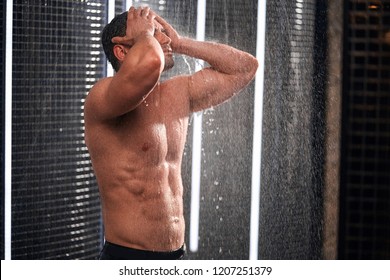 Athletic good looking and attractive man with muscular body standing under running water in shower room with black tiles in background