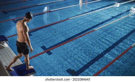 Athletic Male Swimmer Stands On A Starting Block, Ready To Dive Into Swimming Pool. Determined Professional Muscular Athlete Prepares For Championship. High Anglw View.