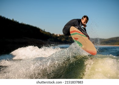 Athletic Male Wakesurfer In Black Wetsuit Jumping On A Wakesurf Board Over The River Waves Against Trees And Blue Sky