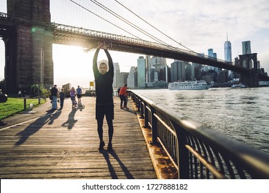 Athletic Man In Black Sportswear Raising Arms Above Head While Stretching On Modern City Waterfront Standing In Bright Back Lit 
