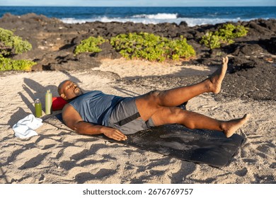 Athletic man performing flutter kicks on an exercise mat during an outdoor workout on a sunny tropical beach with the ocean in the background.