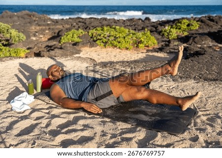 Athletic man performing flutter kicks on an exercise mat during an outdoor workout on a sunny tropical beach with the ocean in the background.