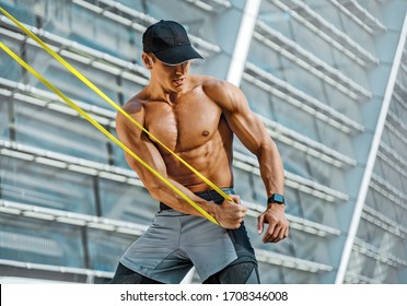 Athletic Man Performs Exercises Using Resistance Band. Photo Of Strong Man Training In The City. Strength And Motivation