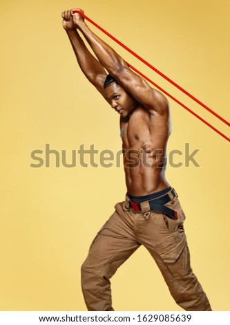 Athletic man performs exercises using resistance band. Photo of african american man on yellow background. Strength and motivation