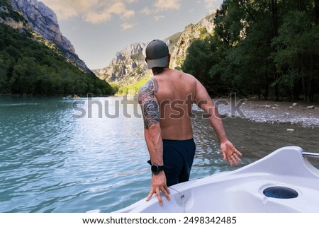An athletic man with a Polynesian tattoo pulls a boat along the water on the Verdon River in southern France on a summer day.