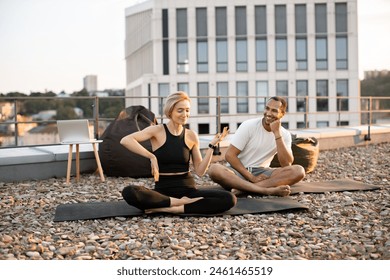 Athletic man and slim woman sitting cross-legged on mat during morning warm-up. Young happy couple having fun dancing after yoga class on roof of modern house. - Powered by Shutterstock