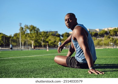 Athletic man soccer player wear blue athletic tank top and black shorts sitting on grassy sports football field, resting after game or workout, look at camera. Take break, sports, lifestyle - Powered by Shutterstock