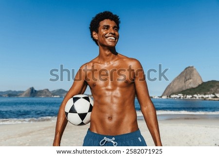 An athletic man stands on the sandy shores of Praia do Flamengo, holding a soccer ball. The famous Sugarloaf Mountain looms in the background against a vibrant blue sky on a perfect beach day.