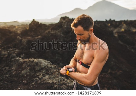 Athletic man start running program on smart watch or fitness tracker. Training topless and using chest heart rate monitor. Trail running outdoors.