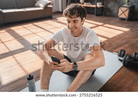 Athletic man using app on mobile phone during home workout, checking results on smartphone after training exercising, sitting on yoga mat, having a rest. Healthy habits lifestyle and gadgets concept.